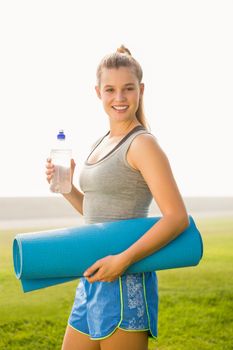 Portrait of sporty blonde holding exercise mat and water bottle in parkland
