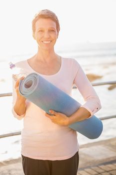 Portrait of smiling sporty woman with exercise mat at promenade