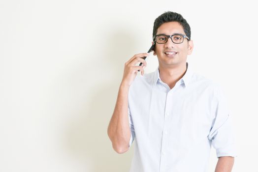 Portrait of handsome casual business Indian man talking on smartphone, standing on plain background with shadow, copy space at side.