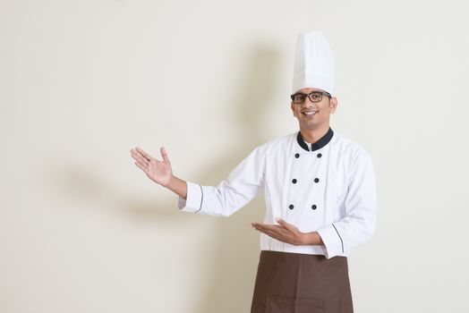 Portrait of handsome Indian male chef in uniform hands gesturing showing something and smiling, standing on plain background with shadow, copy space at side.