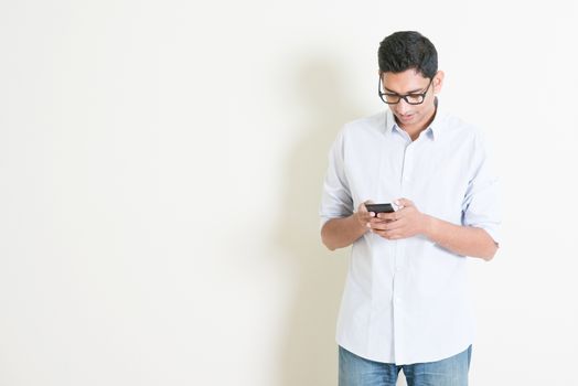 Portrait of handsome casual business Indian man using smartphone, mobile apps concept, standing on plain background with shadow, copy space at side.