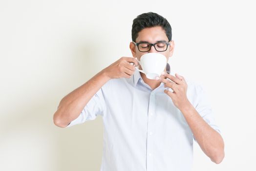 Portrait of handsome casual business Indian guy relax and drinking a cup hot coffee, standing on plain background with shadow, copy space at side.