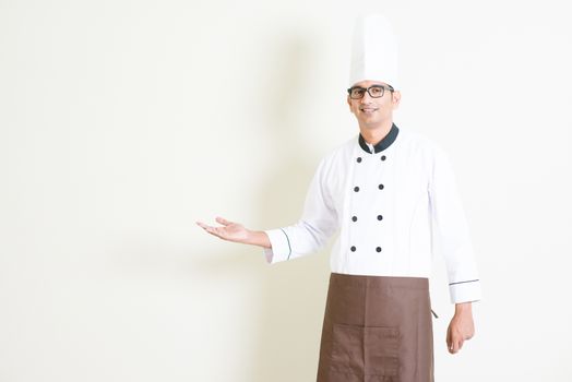 Portrait of handsome Indian male chef in uniform welcoming and smiling, standing on plain background with shadow, copy space on side.