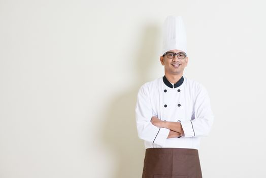 Portrait of handsome Indian male chef in uniform smiling, standing on plain background with shadow, copy space at side.