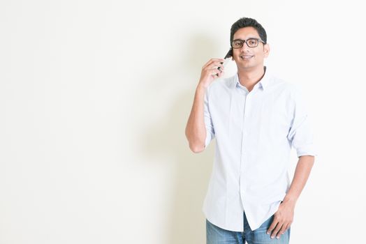Portrait of handsome casual business Indian man using smartphone, standing on plain background with shadow, copy space at side.