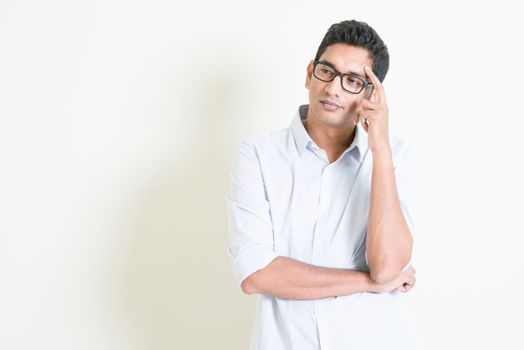 Portrait of handsome casual business Indian man thinking with serious face expression, standing on plain background with shadow, copy space at side.