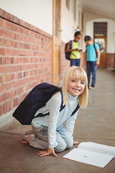 Portrait of cute pupil kneeling over notepad at corridor in school