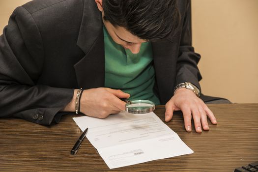 Young Man Sitting at Desk Scrutinizing Paper Contract with Magnifying Glass Prior to Signing