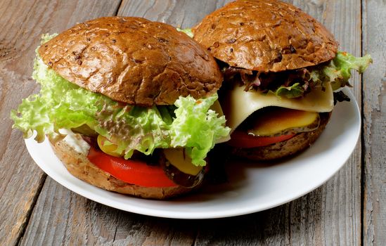 Homemade Hamburgers with Roasted Beef, Lettuce,Tomato, Onions, Cheese, Bacon and Pickle with Whole Wheat Bun on White Plate closeup on Rustic Wooden background