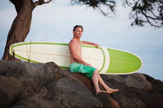 Single adult surfer male outdoors sitting with surfboard on rocks