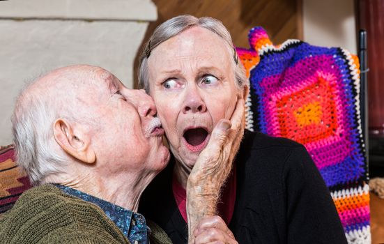 Older gentleman kissing older shocked woman on cheek in livingroom
