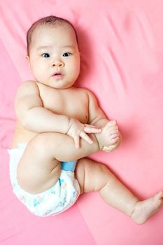 Asian baby girl with diaper touching her foot on pink bed and she looking at the camera