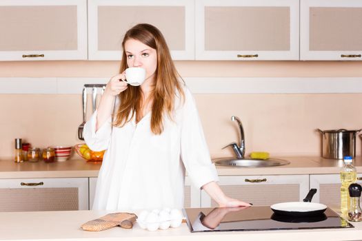 girl in a white men's shirt with long flowing hair is drinking tea in the kitchen in the morning