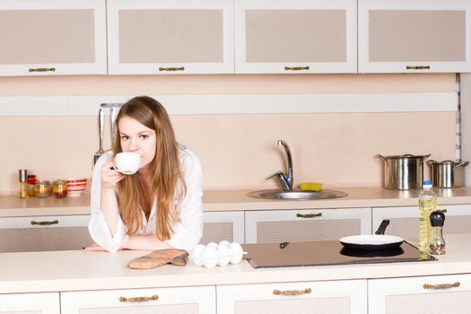 girl in a white men's shirt with long flowing hair is drinking tea in the kitchen elbows on the table in the morning