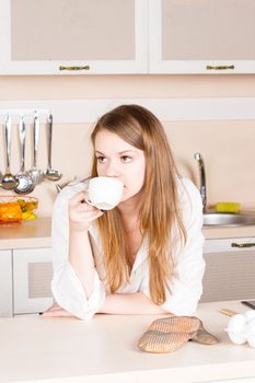 girl in a white men's shirt with long flowing hair is drinking tea in the kitchen elbows on the table in the morning. Vertical framing