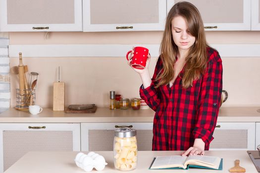 Girl with long flowing hair in a red men's shirt in the kitchen with red cup in his hands. Horizontal framing