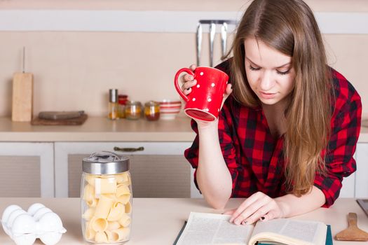 Girl in a red shirt drinking tea and reading in the kitchen. Horizontal framing