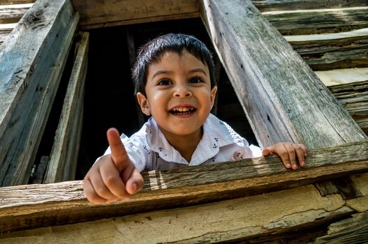 Latino child looking out the window of a wooden house or shed, pointing at the camera and smiling