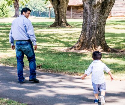 Latino father and child outside walking on a paved path in a park with trees in the background. The child is running after the father.
