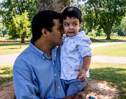 Latino father standing outside in a park with trees behind him holding his son in his arms