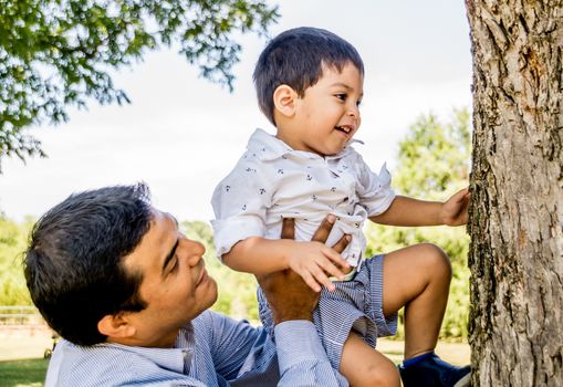 Latino father outside holding his son up to a tree trunk