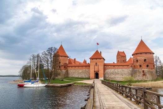 Front view of Trakai Castle with wooden bridge on enterence and small boats in lake, on cloudy sky background.