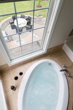 Modern sunken oval bathtub full of clean water in a brown tile surround overlooking a brick patio with outdoor dining through a long view window, viewed from above