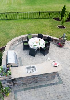 View looking down at a brick paved outdoor living area on an open-air patio with a gas BBQ and concrete kitchen counter alongside a dining table and chairs overlooking green lawns