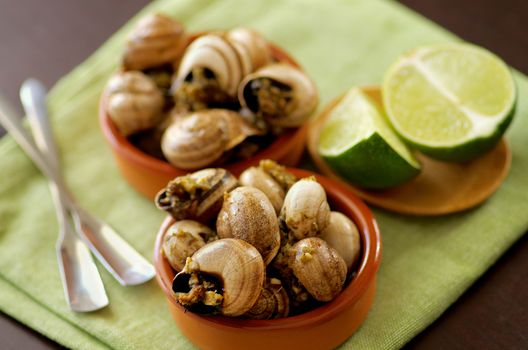 Delicious Escargot with Garlic Butter in Two Bowls with Silver Forks and Sliced Lime closeup on Green Napkin. Focus on Foreground