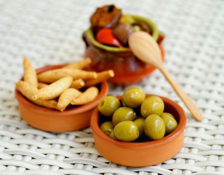 Traditional Spanish Snacks with Green Olives, Bread Sticks and Fried Vegetables in Ceramic Bowls closeup on Wicker background. Focus on Foreground