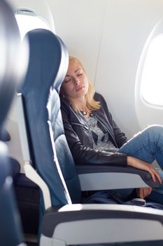 Tired blonde casual caucasian lady napping on uncomfortable seat while traveling by airplane. Commercial transportation by planes.