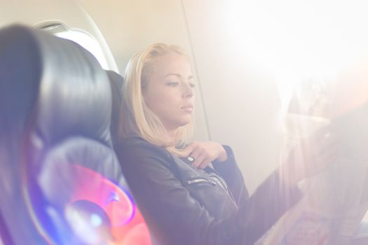 Woman reading newspaper on airplane. Female traveler reading seated in passanger cabin. Sun shining trough airplane window.