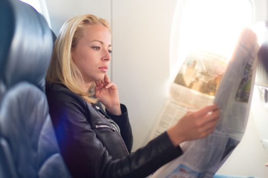 Woman reading newspaper on airplane. Female traveler reading seated in passanger cabin. Sun shining trough airplane window.