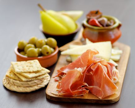 Arrangement of Various Spanish Snacks with Hamon, Goat Cheese, Rosemary Bread Sticks, Green Olives, Fruits and Vegetables closeup on Dark Wooden background. Focus on Foreground