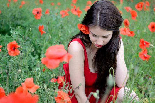 Portrait of shy girl. Female model with red dress surrounded by poppies. Model not facing the camera, head down.