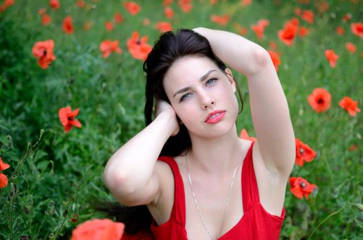 Pretty female model posing with poppies flowers in meadow. Girl putting her hand in hairs.