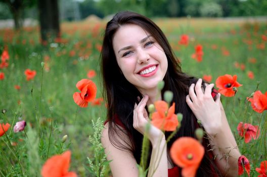 Beautiful polish girl surrounded by field full of poppies. Young female model with charming smile, wearing red dress.