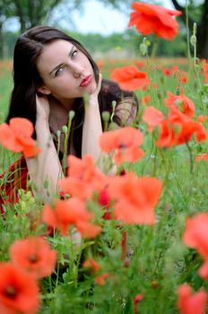 Young female model posing surrounded by poppies flowers. Brunette girl wearing red dress.