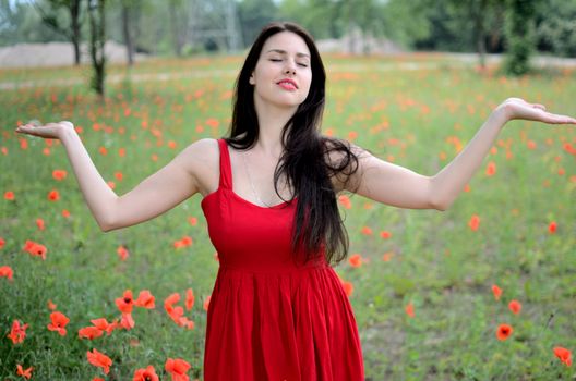 Female model with red dress. Girl meditating in open field, meadow with poppies in background. Model with closed eyes and open arms.