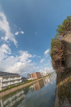 A wide angle shot of a blue sky dotted with clouds reflected in a river flowing below lined with plants on one side and buildings on the other.