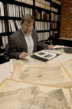 Researcher in archive, searching through maps and photographs. [Maps and photographs on table are public domain. Image on computer creted for this photograph. Model and Property Released.]