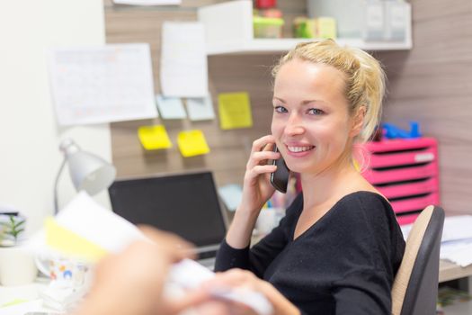 Business and entrepreneurship consept. Beautiful blonde business woman talking on mobile phone in colorful modern creative working environment accepting papers.  Female multi tasking.