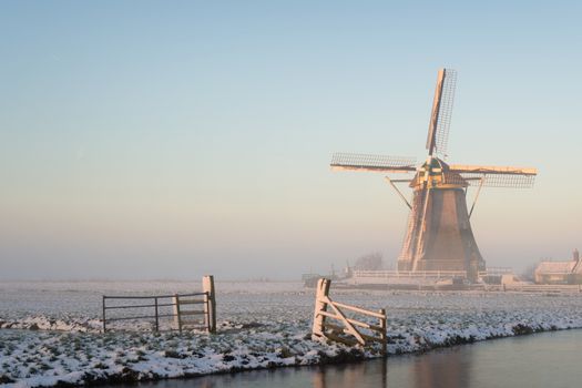 Winter landscape in Holland (the Netherlands) in a rural area with a windmill in a meadow, a fence, a canal, snow and fog at sunrise.