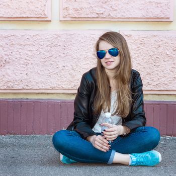 Girl in jacket, jeans and sunglasses sitting on the pavement