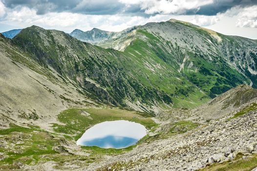 Landscape of Retezat National Park mountains in South Carpatians, Transylvania, Romania, Europe. Small lake with blue sky reflection at center, sheep herd near lake.