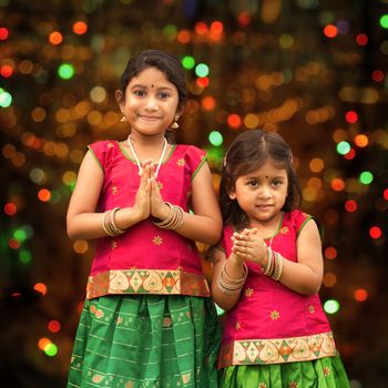 Cute Indian girls dressed in sari with folded hands representing traditional Indian greeting, standing inside a temple celebrating diwali, festival of lights.