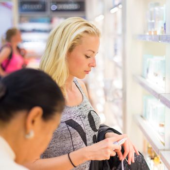 Casual blond young woman smelling perfume in retail store. Beautiful blond lady testing  and buying cosmetics in a beauty store.