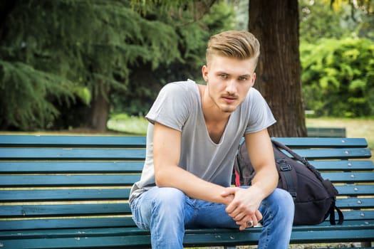 Handsome blond young man sitting on green, wooden park bench