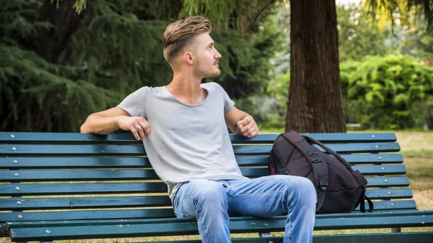 Handsome blond young man sitting on green, wooden park bench