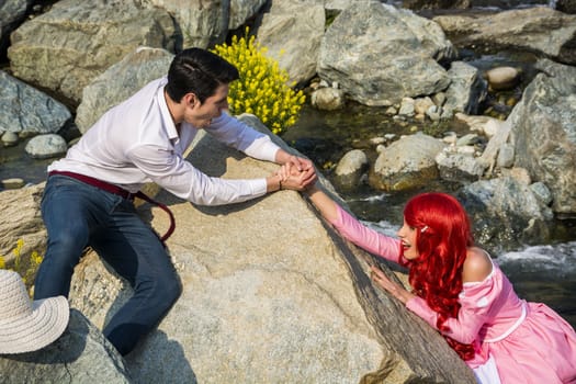 Handsome Young Man Holding on to Hand of Maiden with Bright Red Hair, Helping Woman Falling Over Side of Large Boulder into River